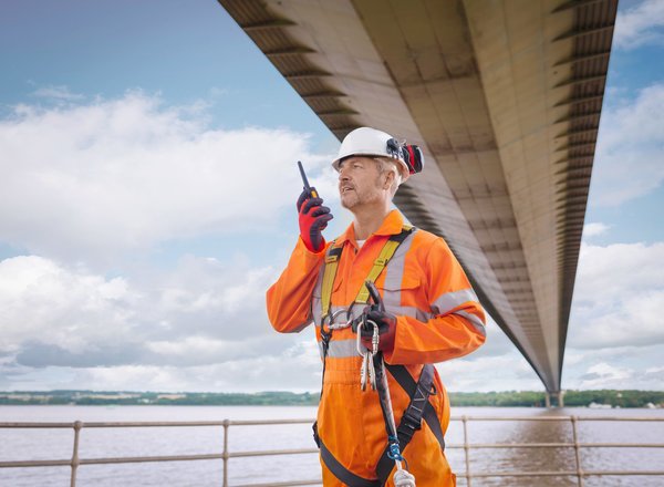 Das Foto ist ein Titelbild der TIPPS, das einen Bauarbeiter mit Persönlicher Schutzausrüstung zeigt, der unter der Humber Bridge an der englischen Ostküste steht und in ein Funkgerät spricht.